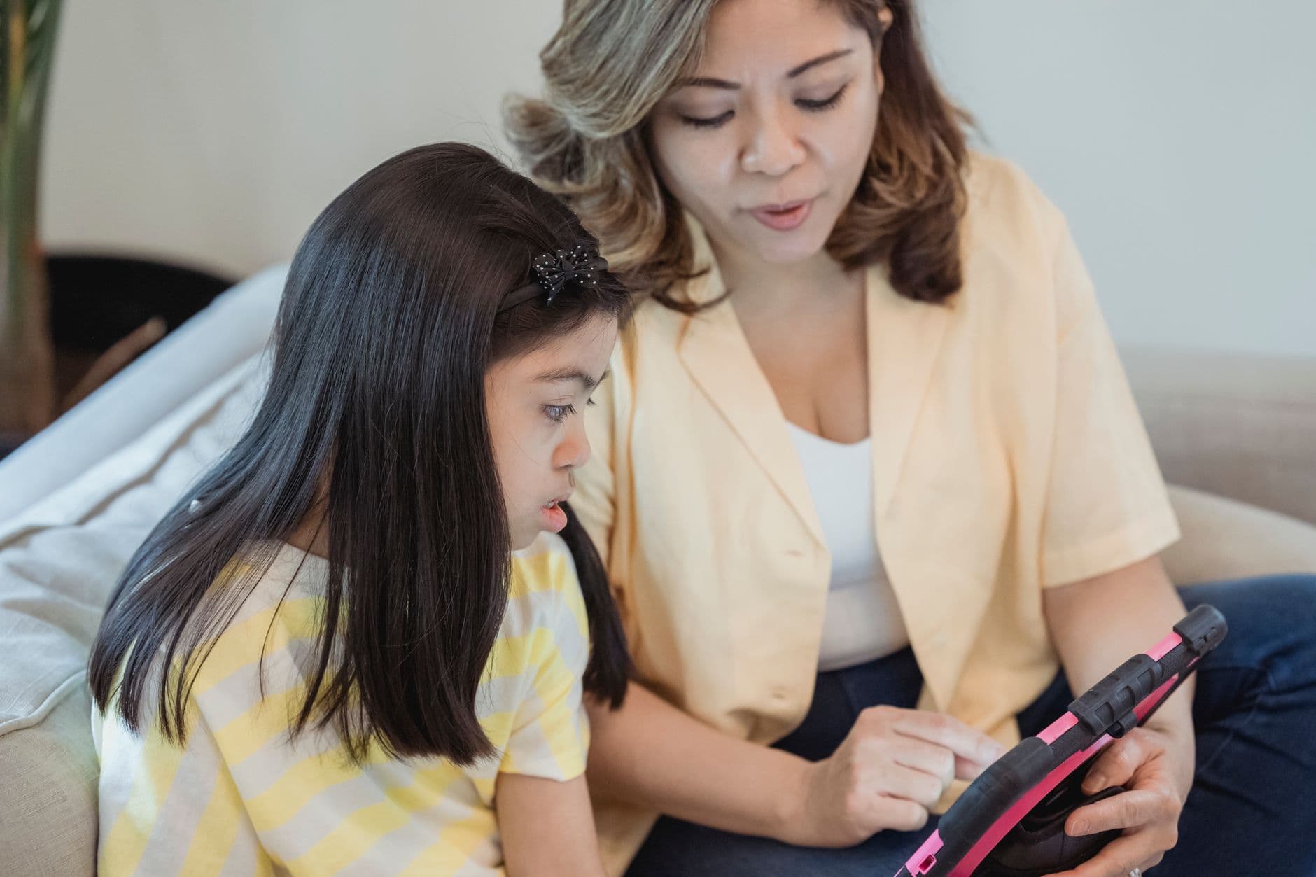 A mother and her young daughter sit together on a couch at home, looking at a tablet
