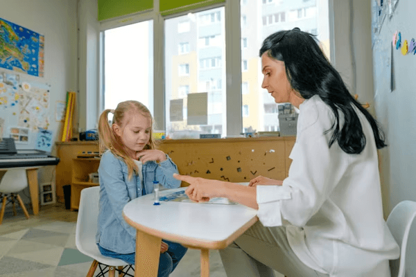 A therapist and a young girl sit at a small table, working on a puzzle activity