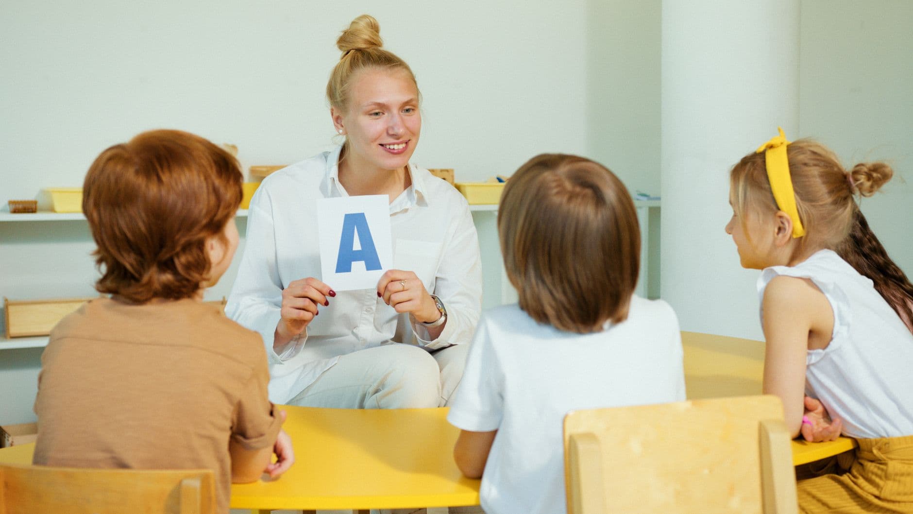 A woman holds up a letter card for three young children sitting around a small yellow table