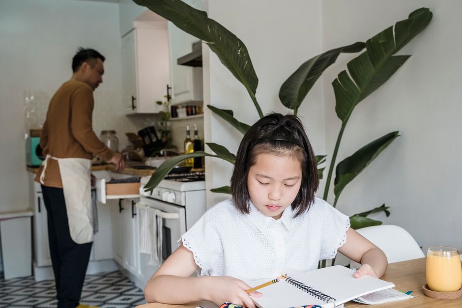 A girl reads her school notebook at the kitchen table while her father cooks behind her