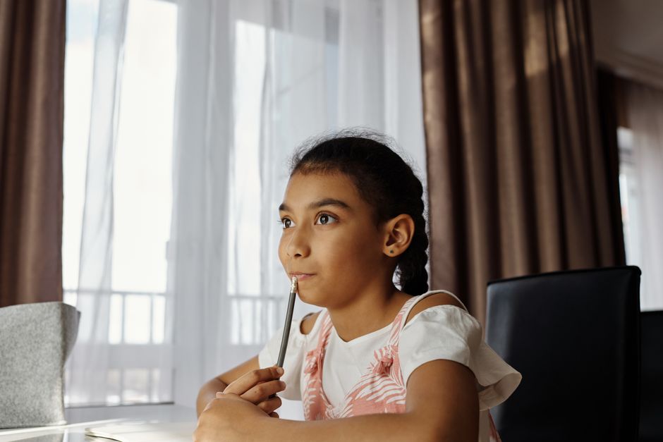 A young girl sitting quietly at a desk with a pencil, looking thoughtful