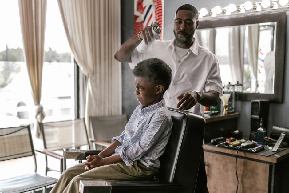 A boy sits in a barber's chair, screwing up his face as the barber sprays his hair